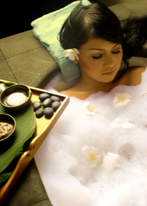 A person lying in a romantic spa bath with rose petals and bubbles, with a tray of candles and glasses of bubbly beside them.